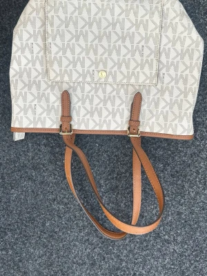 Beige handväska från Calvin Klein - Snygg beige handväska från Calvin Klein med klassiskt CK-mönster över hela väskan. Väskan har bruna läderhandtag, guldfärgade detaljer och en praktisk ficka framtill. Perfekt för dig som gillar stilrena accessoarer med logga.