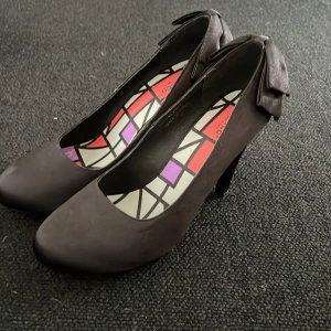 Svarta pumps med rosett - Säljer ett par svarta pumps med hög klack och rund tå. Skorna har en stor dekorativ rosett baktill och insidan har ett färgglatt grafiskt mönster. Perfekta för dig som vill ha något extra till outfiten.