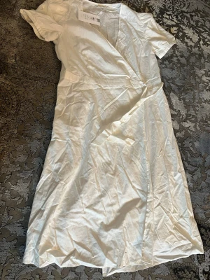 Vit omlottklänning från vila  - Säljer en fräsch vit omlottklänning från vila med korta ärmar och v-ringning. Klänningen har en klassisk omlottdesign och är tillverkad i mjuk bomull, perfekt för varma dagar. Enkel och stilren look som passar till många tillfällen. Klänningen går att stänga och aldrig använts, beställde för många klänningar för förra årets student och inte använt. 