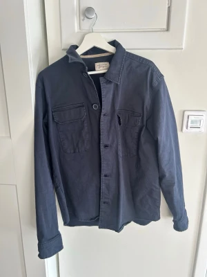 Mörkblå overshirt från Grigio - Stilren mörkblå overshirt från Grigio i bomull. Två bröstfickor med lock och knapp, klassisk krage och knäppning framtill. Perfekt lager-på-lager-plagg med avslappnad passform och snygga detaljer.