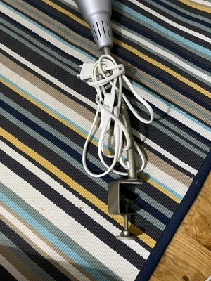 Silver skrivbordslampa med klämfäste - Säljer en silverfärgad skrivbordslampa från IKEA med klämfäste som enkelt kan monteras på bordskanten. Lampan har en rund metallskärm och lång vit sladd med strömbrytare. Perfekt för plugg eller hobbyprojekt.