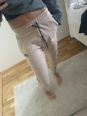 Beige mjukisbyxor från Nike - Säljer ett par beige mjukisbyxor från Nike med svart dragsko i midjan och logga på vänster lår. Byxorna har avsmalnande ben och resår i midjan för skön passform. Perfekta för chill dagar eller träning.