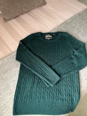 Mörkgrön kabelstickad tröja Superdry - Mörkgrön kabelstickad tröja från Superdry med rund halsringning och långa ärmar. Tröjan har diskreta märkesdetaljer på axeln och vid nederkanten. Perfekt för dig som gillar klassisk stickad stil med modern touch.