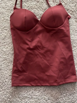 Vinröd body med bygel från Gina Tricot - Säljer en vinröd body från Gina Tricot med bygel och vadderade kupor. Den har smala, justerbara axelband och är tillverkad i ett stretchigt, slätt material som sitter snyggt mot kroppen. Perfekt för att skapa en stilren och trendig look.