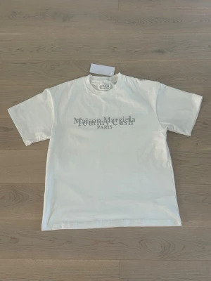 Vit t-shirt från Maison Margiela - Maison Margiela t-shirt i vitt med grått tryck på både fram- och baksidan. Klassisk rund hals och korta ärmar. Materialet är mjuk bomull och passformen är normal. Perfekt för dig som gillar minimalistisk design och exklusiva märken. Är i storlek M men sitter som storlek L!