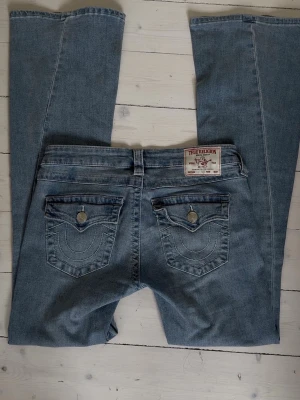 True Religion Joey Low Rise Bootcut Jeans - Säljer ett par ljusblå True Religion Joey jeans med låg midja och flare-ben. Klassisk femficksmodell med snygga detaljer och silverfärgade knappar. Endast använta fåtal gånger.