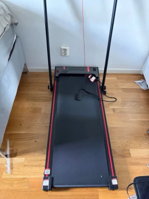 Svart och rött löpband för inomhus - Säljer ett stilrent löpband i svart med röda detaljer. Löpbandet har en digital display och säkerhetslina, samt handtag på båda sidor. Perfekt för hemmaträning och enkel att fälla ihop. Drivs med sladd och har en robust design i metall och plast.