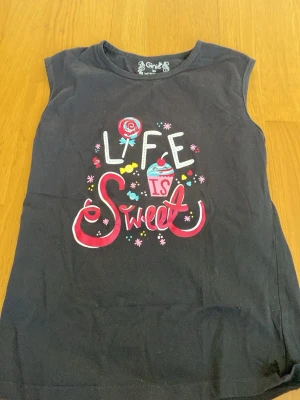 Svart ärmlös topp med tryck från Ginie - Svart ärmlös topp från Ginie med färgglatt tryck på framsidan där det står 'Life is Sweet' tillsammans med godismotiv och cupcake. Toppen har rund halsringning och är gjord i mjukt bomullsmaterial. Perfekt för dig som gillar lekfulla prints.
