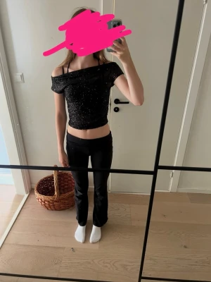 Svart glittrig offshoulder topp - Snygg svart glittrig offshoulder topp med inbyggt linne och som är croppad och har en åtsittande passform. Perfekt för dig som vill sticka ut lite extra med ett trendigt plagg. Toppen är ärmlös och har en glittrig finish som fångar ljuset.