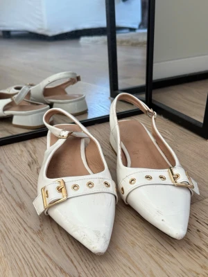 Vita ballerinaskor med gulddetaljer - Säljer ett par vita ballerinaskor med spetsig tå och justerbar rem runt hälen. Skorna har snygga guldfärgade spännen och nitar framtill som ger en trendig look. Materialet är glansigt och insidan är beige.  Tvättar dom extra noga om något är intresserad av dessa! 