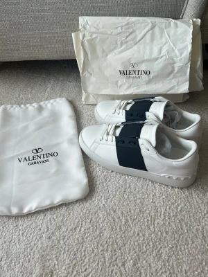 Valentino Garavani vita sneakers - Snygga vita sneakers från Valentino Garavani med bred Mörkblå rem över vristen och vita snören. Skorna är låga och har detaljer i gummi på hälen. Tillverkade i slätt läder och kommer med dustbag och originalpåse.