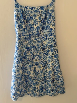 Blåvit blommig sommarklänning - Supersöt blåvit klänning med blommigt mönster och smala axelband. Klänningen är perfekt för varma dagar. Materialet känns svalt och mjukt, och den har en feminin siluett som ger en fräsch look.
