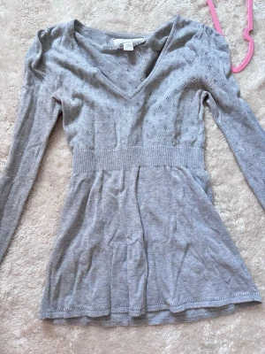 Grå stickad babydoll från American Eagle - Supersöt grå stickad babydoll från American Eagle med v-ringning och lång ärm. Blusen har ett fint hålmönster upptill, markerad midja och lätt utställd nederdel. Perfekt till jeans eller kjol för en chill och snygg look.