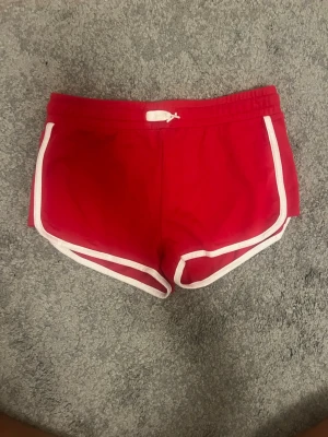 Röda sportiga shorts med vit kant - Snygga röda shorts med vit kant och snörning i midjan. Klassisk sportig look med rundade ben och elastisk midja. Perfekta för sommardagar eller träning. Materialet känns mjukt och lätt, troligen bomull eller polyester.