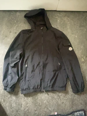 Svart vindjacka från Moncler - Snygg svart vindjacka från Moncler med huva och dragkedja framtill. Jackan har två fickor med dragkedja och en ficka med knapp på ärmen där Moncler-loggan sitter. Tillverkad i lätt och slitstarkt material, perfekt för blåsiga dagar om du vill också du kan vika in luvan så den blir grisch jacka 