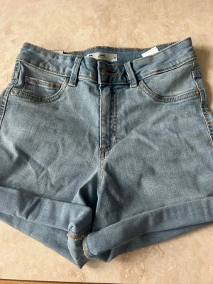 Ljusblå denimshorts - Snygga ljusblå denimshorts från Jane med klassisk femficksdesign och uppvikta benslut. Shortsen har bälteshällor och stängs med knapp och dragkedja. Perfekta för varma dagar och enkel att matcha med olika toppar.