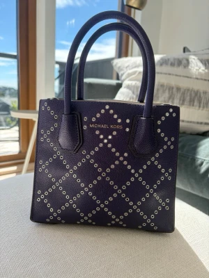 Lila handväska från Michael Kors - Snygg lila handväska från Michael Kors i fyrkantig modell med silverfärgade nitar i rutmönster. Väskan har två handtag och en avtagbar axelrem. Oanvänd och jättebra skick! 