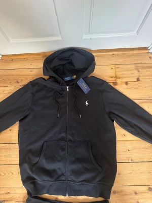 Svart set från Polo Ralph Lauren - Säljer ett svart set från Polo Ralph Lauren med hoodie och byxor. Hoodien har dragkedja, fickor och justerbar huva. Byxorna har dragsko i midjan och fickor. Båda delarna har den klassiska vita Polo-loggan broderad på bröstet och benet. Mjukt och bekvämt material.