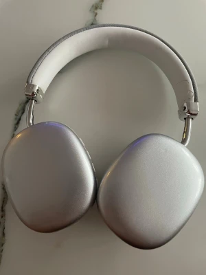 AirPods Max silver - Säljer ett par Apple AirPods Max i silver. Over-ear hörlurar med stilren design, aluminiumkåpor och mjuka mesh-öronkuddar. Huvudbandet är klätt i vitt tyg och öronkuddarna är grå. Perfekt för dig som vill ha grymt ljud och snygg look.