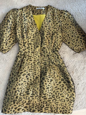 Leopardmönstrad gul klänning Adoore - Säljer en kort klänning från Adoore i glansigt gult tyg med leopardmönster. Klänningen har v-ringning, puffiga korta ärmar och markerad midja. 