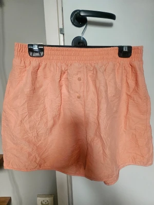 Corallfärgade shorts från H&M - Säljer ett par shorts från H&M med elastisk midja och två knappar framtill. Shortsen har en avslappnad passform och är tillverkade i ett lätt och skönt material, perfekta för varma dagar.