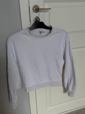 Vit  sweatshirt - En enkel vit croppad sweatshirt med rund hals och långa ärmar. Tröjan har ribbade muddar vid ärmslut och nederkant. Perfekt till jeans eller mjukisbyxor för en avslappnad look. Tillverkad i mjukt bomullsmaterial.