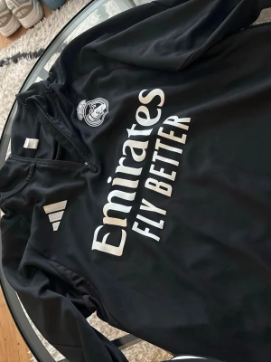 Svart Real Madrid träningströja Adidas - Säljer en svart långärmad träningströja från Adidas med Real Madrid-logga och Emirates Fly Better-tryck på bröstet. Tröjan har halv dragkedja framtill och vita detaljer. Perfekt för fotbollsträning eller gymmet.