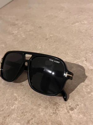 Svarta solglasögon från Tom Ford - Snygga svarta solglasögon från Tom Ford med markerad båge och gulddetaljer vid skalmarna. Glasen är mörka och modellen har en modern, fyrkantig form som ger en cool och exklusiv look. Perfekta för dig som vill sticka ut med stil.