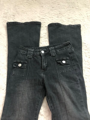 Svarta utsvängda jeans med fickor - Säljer ett par svarta jeans med utsvängda ben och dekorativa fickor framtill med silverknappar. Jeansen har hög midja och markerade sömmar längs benen. Perfekta för dig som gillar retrostil och vill ha något som sticker ut.