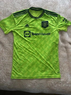 Grön tröja  - Säljer en neongul Manchester United målvaktströja från Adidas med svarta detaljer och TeamViewer-logga på bröstet. Tröjan har korta ärmar, rund hals och ett mönster i ljusare grönt. Perfekt för fotbollsträning eller match.