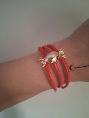Rött armband med godisdetalj - Snyggt armband i rött konstläder som lindas tre varv runt handleden. Armbandet har en guldfärgad detalj i form av ett godis i mitten som ger en lekfull och unik look. Perfekt accessoar för dig som gillar färg och roliga detaljer.