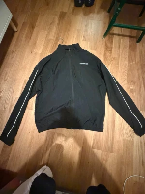 Svart vindjacka från Reebok - Svart vindjacka från Reebok med dragkedja framtill och vit piping längs ärmarna. Jackan har hög krage och Reebok-logga på bröstet. Perfekt för blåsiga dagar och sportiga outfits.