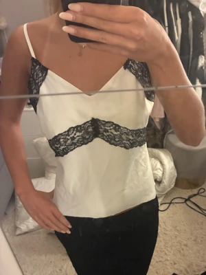 Vit linnetopp med svarta spetsdetaljer - Snygg vit linnetopp med tunna axelband och svarta spetsdetaljer vid byst och midja. Toppen har en v-ringad halsringning och är perfekt för en trendig look. Materialet känns mjukt och lätt mot huden.