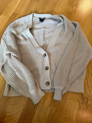 Beige stickad kofta med knappar - Mysig beige stickad kofta med tre stora knappar framtill och bred ribbad kant. Koftan har en avslappnad passform, djup v-ringning och långa ärmar med ribbade muddar. Perfekt att slänga över en t-shirt eller linne för en chill look.