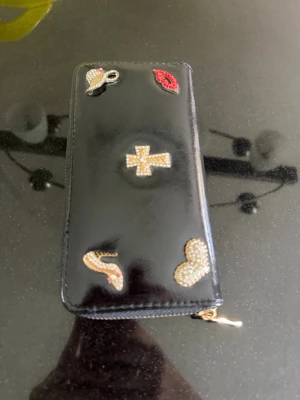 Svart plånbok med blingdetaljer - Svart plånbok i glansigt material med dragkedja runtom. Framsidan är dekorerad med blingade detaljer. Perfekt för dig som gillar unika accessoarer med lite extra bling.