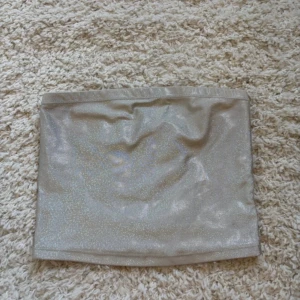Glittrig silver tubtopp - Snygg glittrig tubtopp i silver med skimrande yta. Perfekt för dig som vill sticka ut och addera lite extra bling till din outfit. Axelbandslös och croppad modell som sitter tight runt överkroppen.