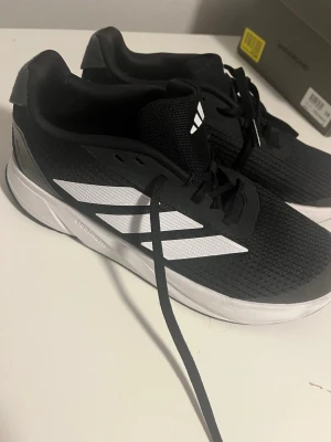 Svarta Adidas lightmotion - Snygga svarta sneakers från Adidas med klassiska vita ränder på sidan. Skorna har en låg profil, svart mesh-överdel och vit sula. Perfekta för en sportig och avslappnad stil. Lätta och bekväma med snörning framtill. 