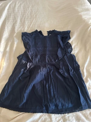 Mörkblå blus med volanger Holly & Whyte - Supersöt mörkblå blus från Holly & Whyte by Lindex med volangdetaljer längs ärmarna och bröstet. Blusen har rundad hals med liten volangkant och fina spetsinslag framtill. Tillverkad i ett lätt och luftigt tyg, perfekt för en trendig look.