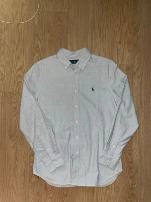 Vit skjorta från Ralph Lauren - Klassisk vit skjorta från Ralph Lauren med blå broderad logga på bröstet. Skjortan har lång ärm, knappar framtill och button-down krage. Tillverkad i mjuk bomull för en clean och tidlös look.
