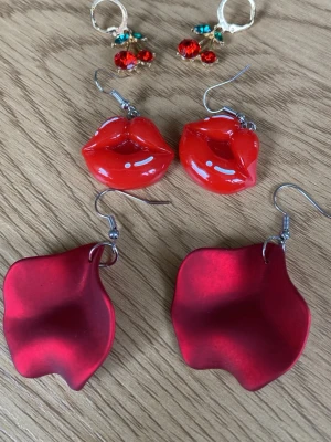 Tre par röda statementörhängen - Säljer tre par unika örhängen: ett par med röda läppar i plast, ett par formade som röda blomblad i matt material och ett par guldfärgade hoops med röda och turkosa pärlor. Perfekta för dig som vill sticka ut med färgglada accessoarer.