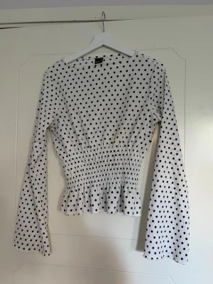 Vit prickig topp med smock - Supersöt vit topp med svarta prickar och smockad midja som ger en snygg siluett. Toppen har långa ärmar och lätt volang nertill. Perfekt för dig som gillar trendiga detaljer och ett lekfullt mönster.