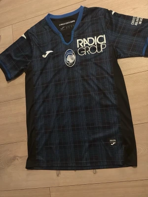 Atalanta matchtröja Holm #3 Joma - Säljer en officiell Atalanta Bergamo fotbollströja från Joma, med spelarnamn Holm och nummer 3 på ryggen. Tröjan har ett blått och svart rutigt mönster, korta ärmar och tryck för Christmas Match 2023. Materialet är lätt och andas, perfekt för match eller träning.