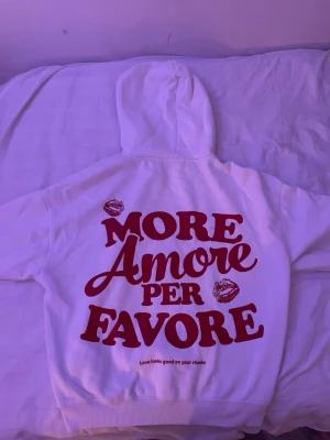 Vit hoodie med röd text och hjärta - Vit hoodie med stor röd text 'More Amore Per Favore' och kyssmärken på ryggen. På framsidan finns en liten röd hjärtdetalj vid halsen och en klassisk magficka. Hoodien har huva och ribbade muddar. Perfekt för dig som gillar statement-plagg och romantiska detaljer.