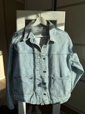 Ljusblå jeansjacka från ZARA - Snygg ljusblå jeansjacka från ZARA med klassisk krage, stora framfickor och knäppning framtill. Jackan har en avslappnad passform och är tillverkad i slitstarkt denimtyg. Perfekt för dig som vill ha en tidlös och trendig look.