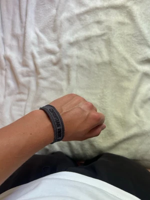 Grått tygarmband från Christian Dior - Snyggt grått armband från Christian Dior i vävt tygmaterial med svart logotyptext. Armbandet har knytning med tofsar i ändarna och en stilren, avslappnad look som passar perfekt till streetwear.