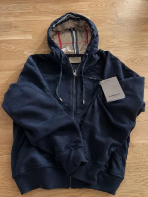 Svart hoodie från Burberry - Snygg svart hoodie från Burberry med dragkedja och klassiskt rutigt foder i huvan. Hoodien har ribbade muddar, fickor framtill och justerbar dragsko i huvan. Perfekt för dig som vill ha en stilren och exklusiv look.