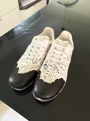 Maison Margiela svartvita sneakers - Maison Margiela sneakers i vitt läder med svart tåparti och svart färgstänk över hela ovandelen. Klassisk låg modell med vita skosnören och gummisula. Unik design som sticker ut och passar perfekt för dig som gillar statement-plagg.