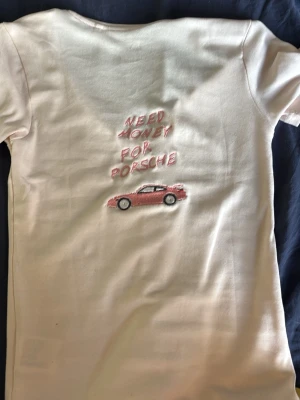 Vit t-shirt med broderad Porsche-text - Säljer en vit t-shirt med djup v-ringning och korta ärmar. På ryggen finns ett broderat motiv med texten 'NEED MONEY FOR PORSCHE' och en liten rosa bil. T-shirten är figurnära och gjord i mjuk bomull, perfekt för en avslappnad och trendig look. En unik tröja gjord av mig 🌸!!! Om ni har annan storlek, skriv till mig så kan det fixas💗 samt om ni vill ha en annan färg