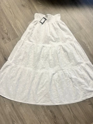 Vit långkjol från Blossom - Luftig vit långkjol från Blossom med smock i midjan och diskret blommigt mönster. Kjolen har volangdetaljer och är perfekt för varma dagar. Tillverkad i ett lätt och bekvämt material.