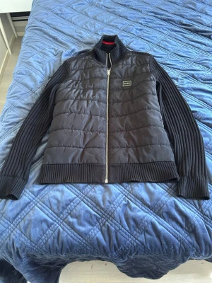 Svart stickad tröja från Hugo Boss - Snygg svart stickad tröja från Hugo Boss med quiltad framsida och ribbstickade ärmar och rygg. Tröjan har dragkedja framtill, hög krage och en diskret logotyp på bröstet. Perfekt för dig som gillar stilrena och moderna plagg. Köpt för 1999kr och jag säljer den för 699. Taggarna här tyvärr åkt av och jag har klippt bort en av dem. Pris kan diskuteras.
